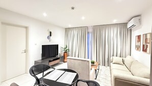 A modern living room with a flat-screen TV, a sofa, a dining table with chairs, and a large window with curtains. - Serene One- Luxury 1BR - The Bantree Accra Airport (Accra)