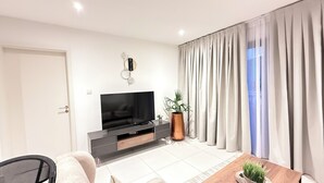 A modern living room with a flat-screen TV, a coffee table, a sofa, a potted plant, and a large window with curtains. - Serene One- Luxury 1BR - The Bantree Accra Airport (Accra)