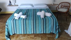 Ein Zimmer mit einem Bett, zwei Nachttischen, einem Korbstuhl, einer Hängeleuchte und einem Gemälde einer Küstenlandschaft.