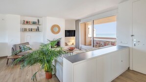 Ein modernes Wohnzimmer mit einer Couch, einem Couchtisch und einer großen Topfpflanze. Es gibt eine Essecke mit Tisch und Stühlen sowie Meerblick durch ein großes Fenster.