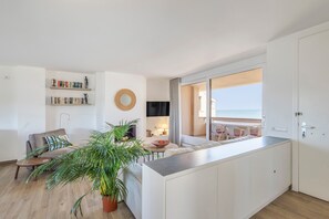 A modern living room with a sofa, coffee table, and a large potted plant. There is a dining area with a table and chairs, and a view of the sea through a large window. - Holiday Home 'Frontline Apartment with Community' with Sea View, Shared Pool and Wi-Fi (Pals)