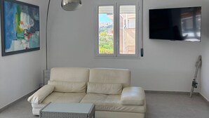 A living room with a sofa, a coffee table, a painting, a TV, and a window with a view.