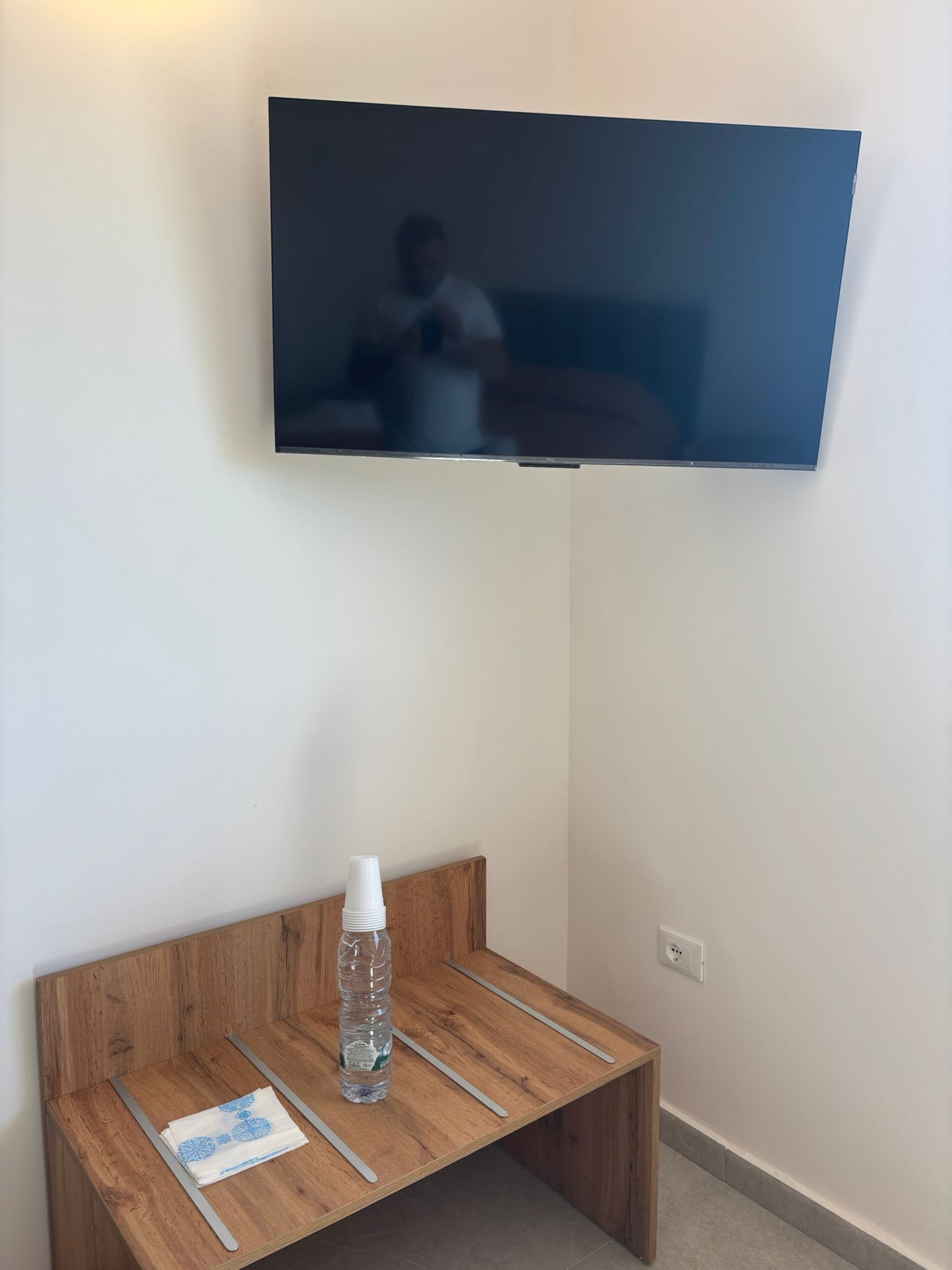 A TV mounted on a wall, a wooden table with a water bottle and a folded towel, and a power outlet on the wall.