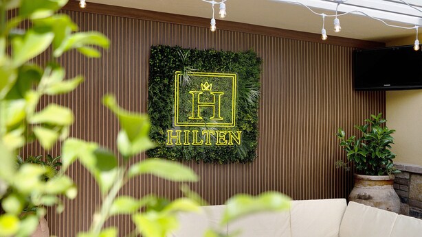 Een muur met een logo en het woord 'HILTEN', in een kamer met een bank en een plant in pot.