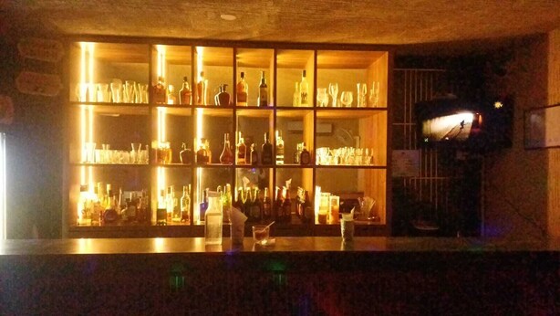 A bar with shelves of bottles and glasses, a counter with a drink, and a view of a building outside. - Hotel Tamilnadu Trichy (Tiruchirappalli)