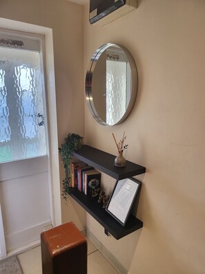 A room with a door, a round mirror, a black shelf with books and a small vase, and a brown stool. - 3-Bed Detached Home. Contractors & Families. Quiet cul-de-sac. Parking. (Wolverhampton)