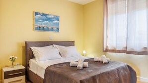 Een keurig opgemaakt bed met witte lakens, een nachtkastje met een bloevas, en een schilderij van een strandlandschap aan de muur.