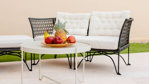 Een ronde tafel met een fruitschaal, twee rieten stoelen en een witte, gestoffeerde bank.