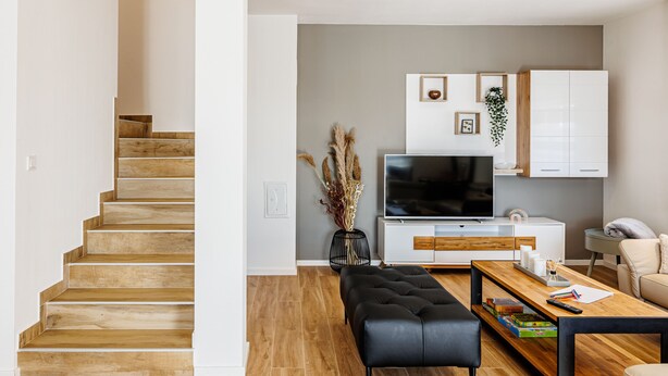 Een moderne woonkamer met een trap, houten vloeren, een zwart leren poefje, een houten salontafel, een televisie en een witte kast.