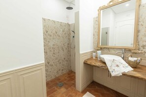 Uma casa de banho com chuveiro, uma pia com bancada de madeira, um espelho com moldura ornamentada e um papel de parede com padrão floral.