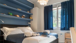 A bedroom with a bed, a nightstand, a chair, a window with curtains, and a built-in shelf with books. - CASA CONCETTA - View with a balcony and a large window (Rapallo)
