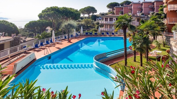 A swimming pool with a clear blue water, surrounded by palm trees and a view of residential buildings. - CASA CONCETTA - View with a balcony and a large window (Rapallo)
