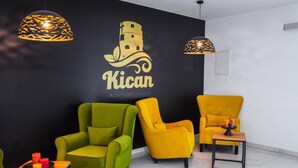 Eine Lobby mit schwarzer Wand, dem Kican-Logo, grünen und gelben Stühlen, einem kleinen Tisch mit Kerzen und Pendelleuchten.