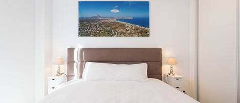 Habitación de hotel con cama, dos mesitas de noche con lámparas, un ventanal grande y un cuadro de un paisaje.