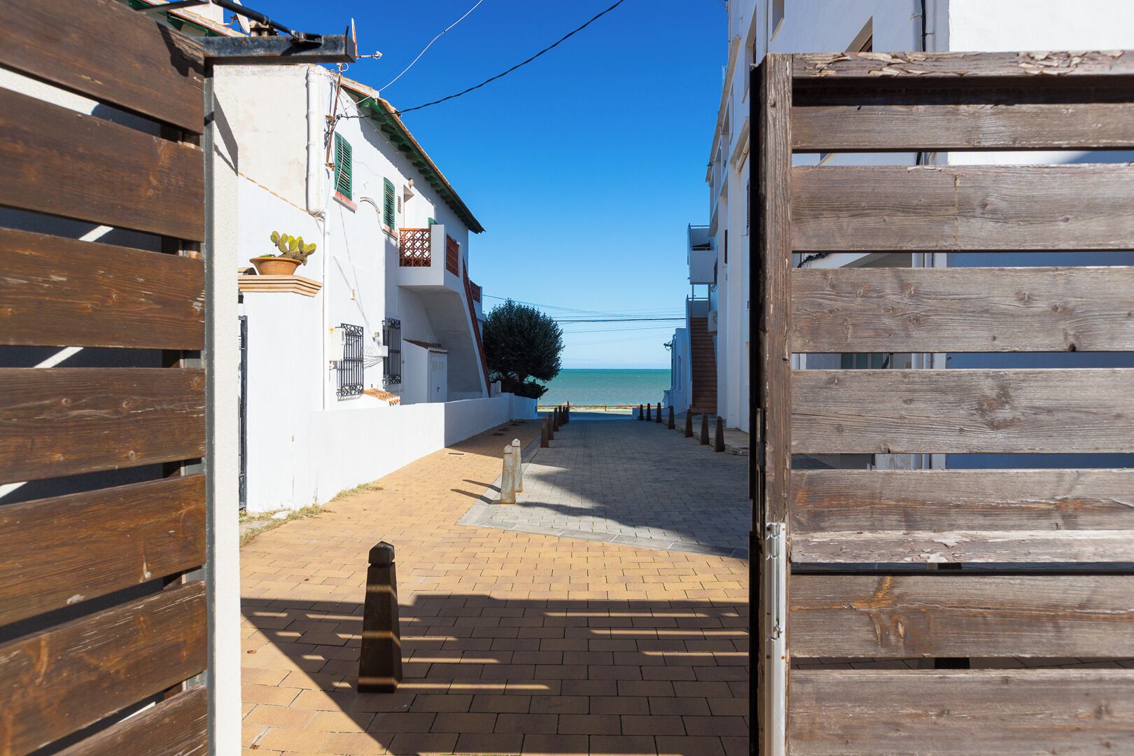 A narrow paved street leading to a beach, flanked by white buildings with balconies and wooden gates.