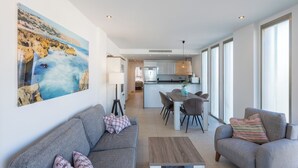 A modern living room with a sofa, a coffee table, and a dining area with chairs. There is a kitchen in the background with a fridge and a hanging light fixture. - Beachfront Premium Terraza piscina Javea (Javea)
