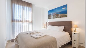 A neatly made bed with a headboard, a bedside table with a lamp, and a window with curtains. - Beachfront Premium Terraza piscina Javea (Javea)