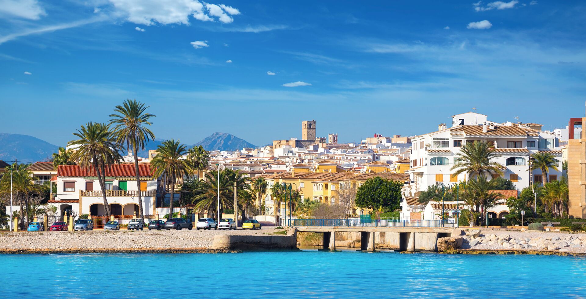 A coastal cityscape with buildings, palm trees, and a bridge over water.
