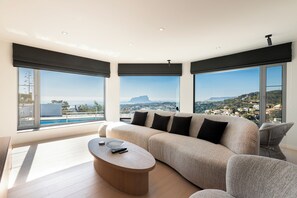 A modern living room with a large sofa, a coffee table, and large windows offering a city view. - Villa Rayo del Sol (Benissa)