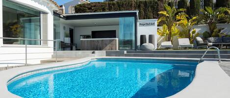 A modern pool area with a clear blue pool, a lounge with sun loungers, and a building with large windows.