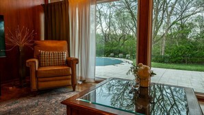 A room with a leather armchair, a glass coffee table, and a view of a swimming pool and greenery.