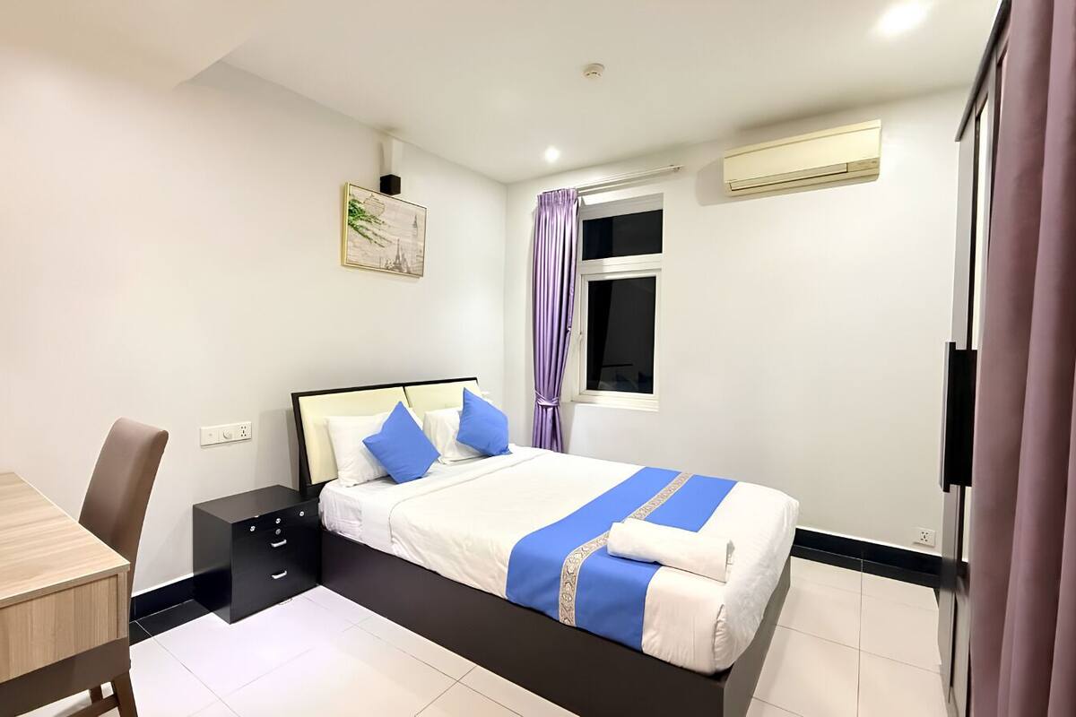 Un dormitorio con cama, escritorio, silla, mesita de noche, ventana con cortinas y unidad de aire acondicionado.