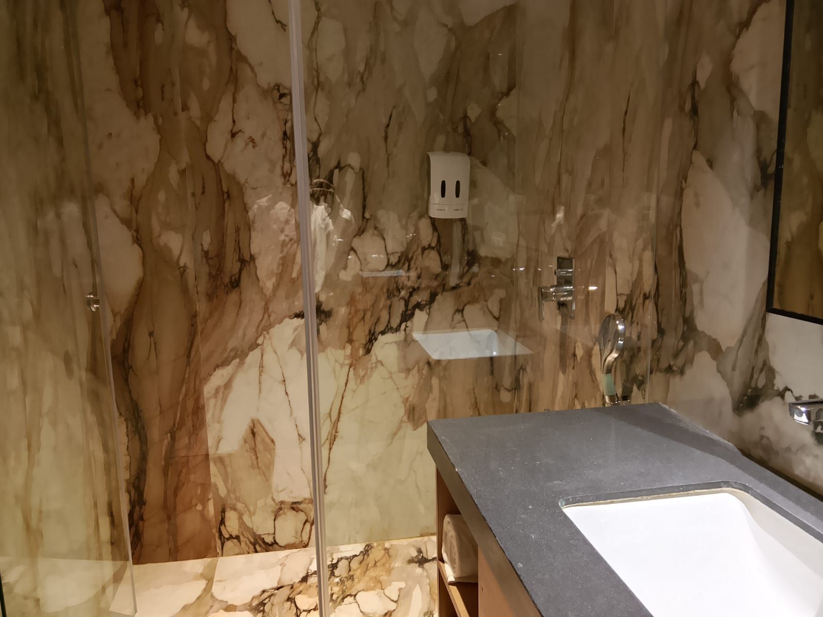Un baño con pared de mármol, cabina de ducha de vidrio, lavabo y espejo.