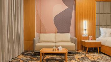 Una habitación de hotel con un sofá, una mesita con un tarro de cristal y una tarjeta, una mesita de noche con botellas, y una alfombra estampada.