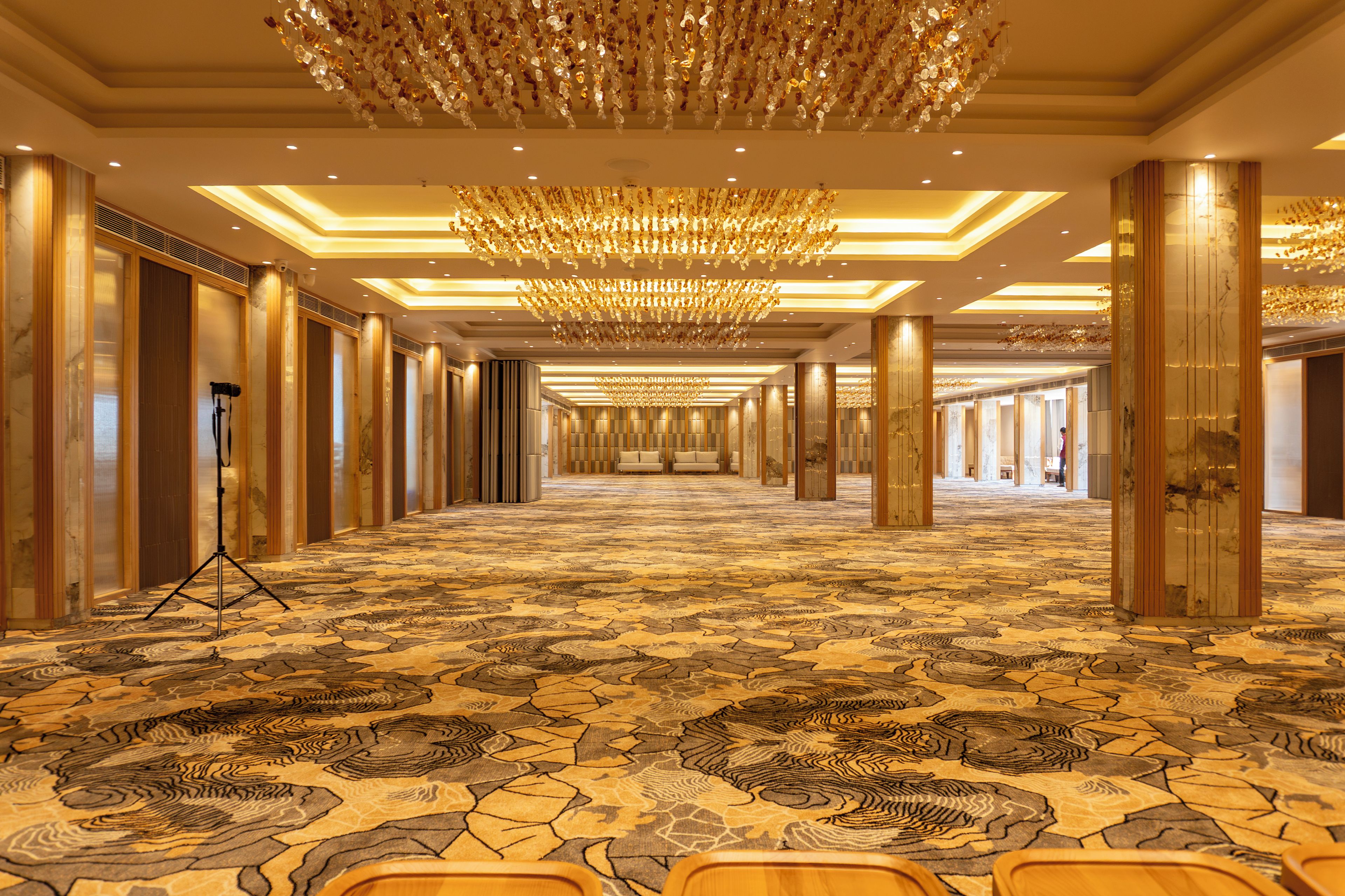 A hotel corridor with patterned carpet, a chandelier, and a camera tripod.