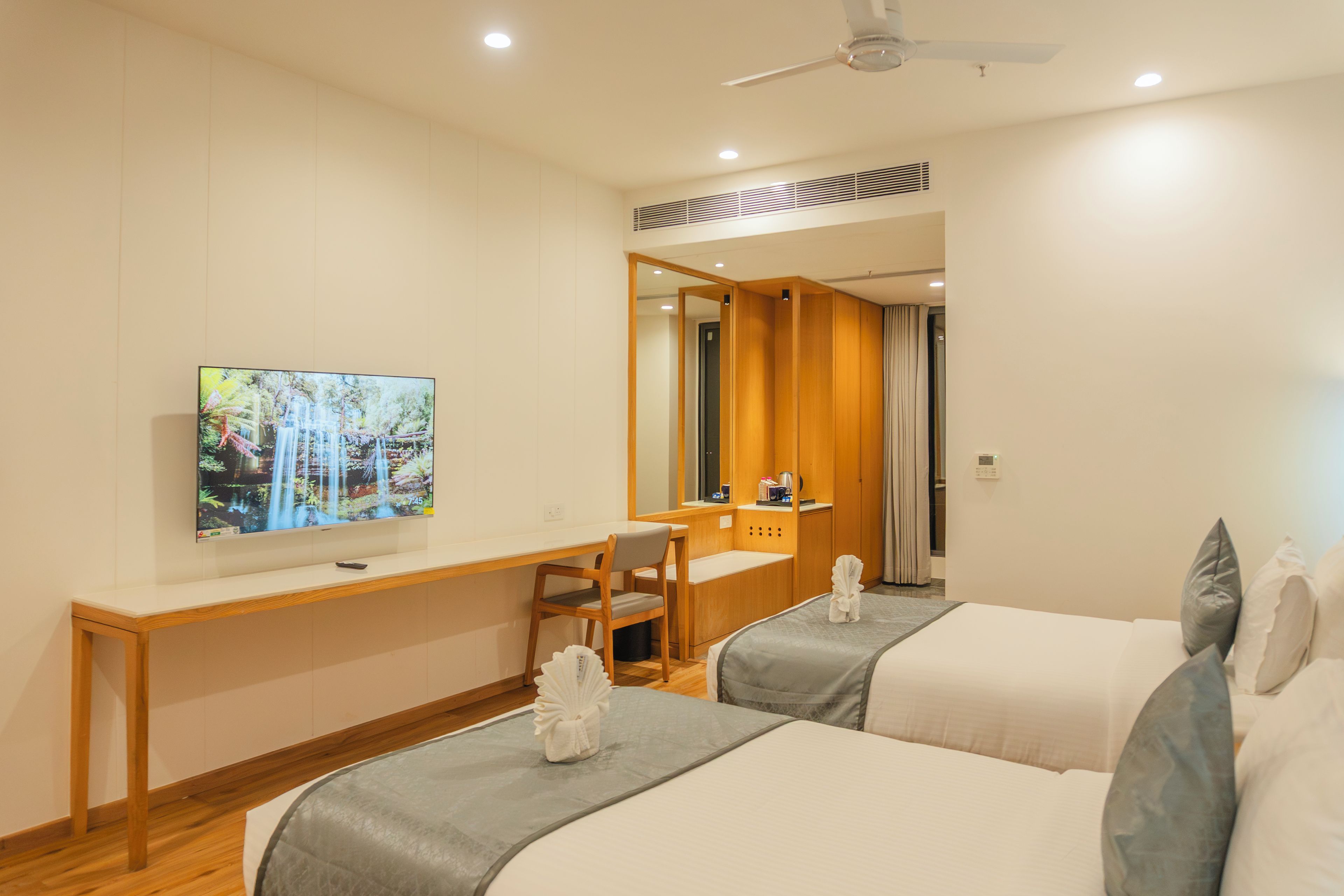 A hotel room with two beds, a desk, a television, and a ceiling fan.
