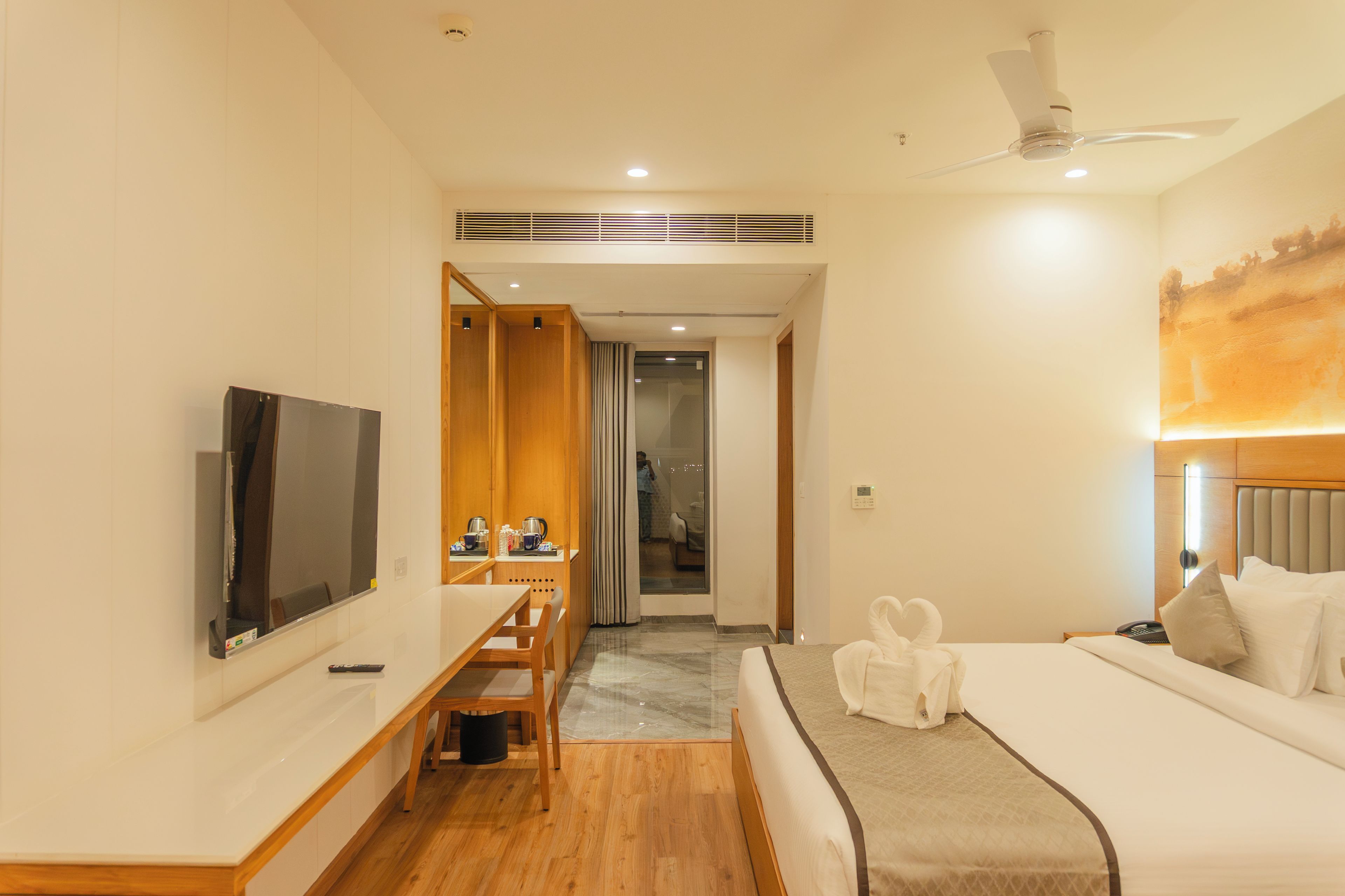 Ein Hotelzimmer mit einem Bett, einem Fernseher, einem Schreibtisch mit Stuhl, einem durch eine Glastür einsehbaren Badezimmer und einem Deckenventilator.