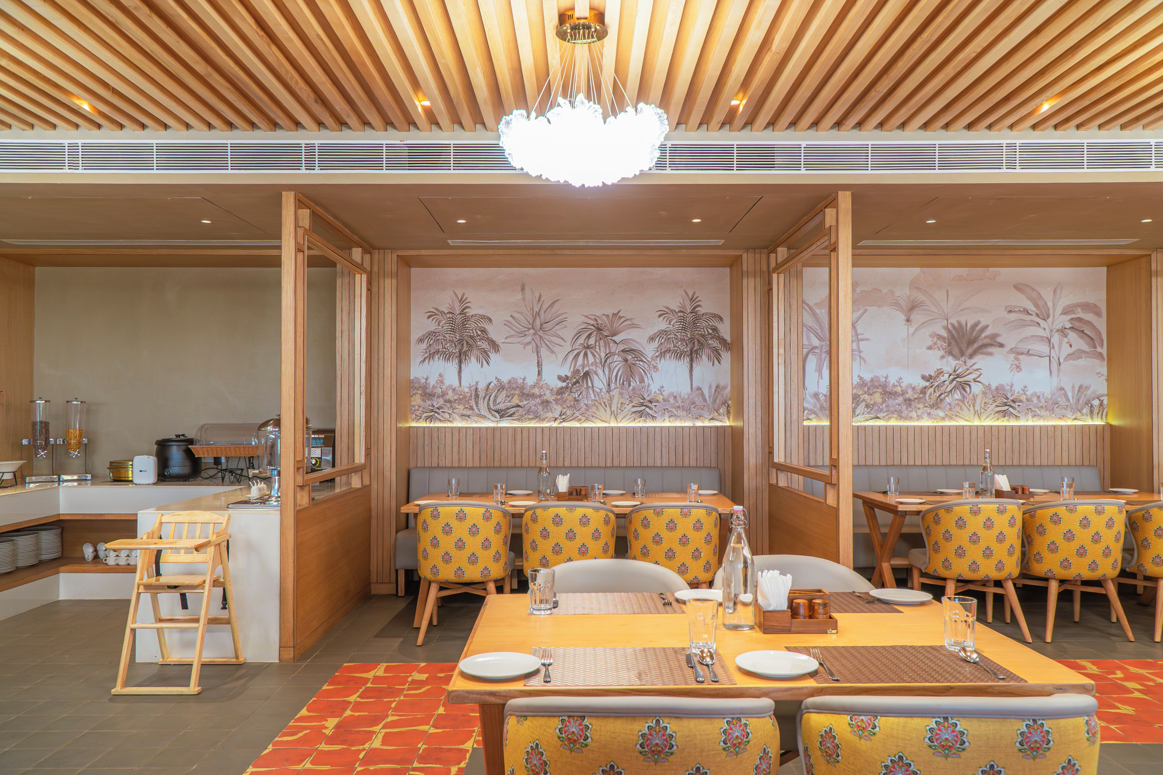 A restaurant with wooden ceiling beams, a central chandelier, and a mural of tropical plants on the back wall.