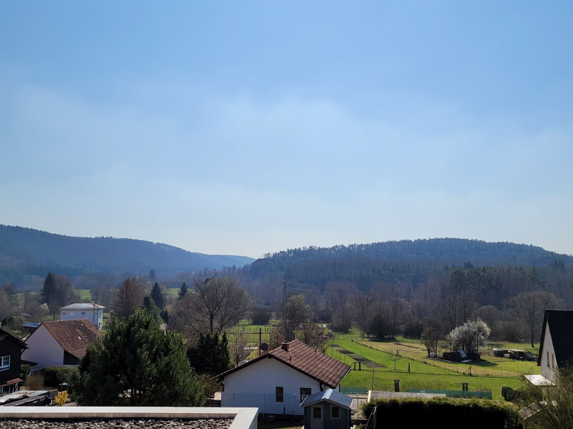 Ländliche Landschaft mit Häusern, blauem Himmel und sanften Hügeln im Hintergrund.