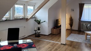 Un espacio habitable moderno estilo loft con techo inclinado, piso de madera, comedor con mesa y sillas, televisor sobre un mueble de madera y un ventanal con vistas a árboles.