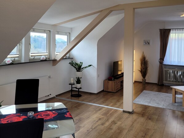 Un espacio habitable moderno estilo loft con techo inclinado, piso de madera, comedor con mesa y sillas, televisor sobre un mueble de madera y un ventanal con vistas a árboles.