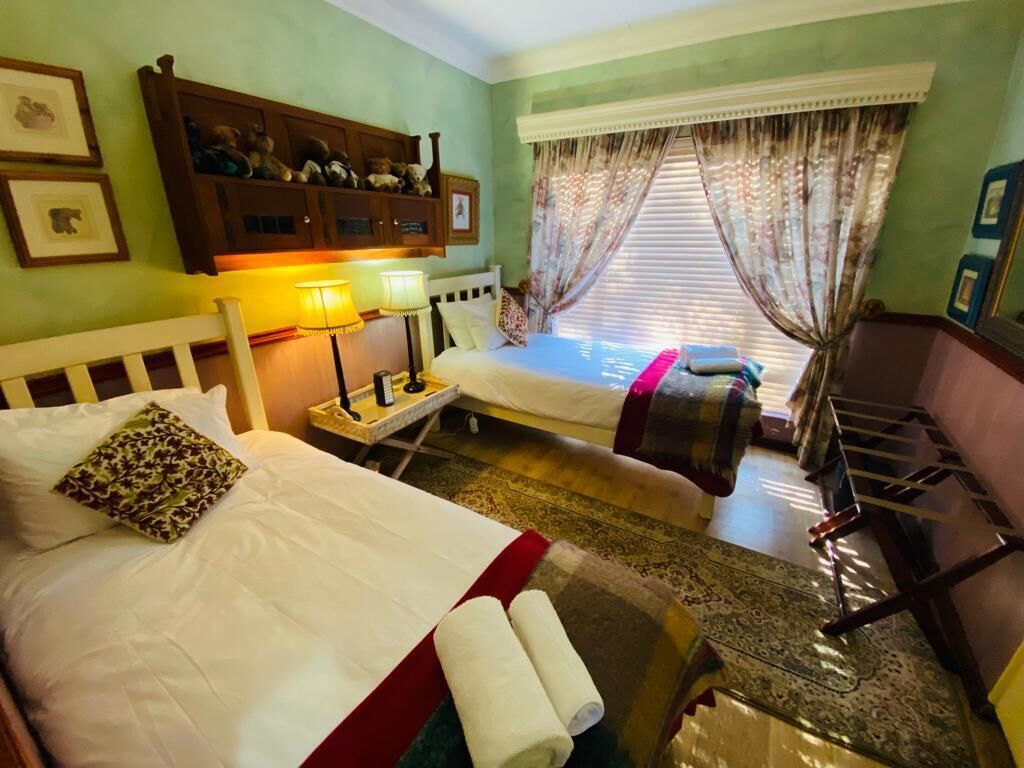 A hotel room with two beds, a nightstand with a lamp, a wooden headboard, a window with curtains, and a shelf with decorative items.