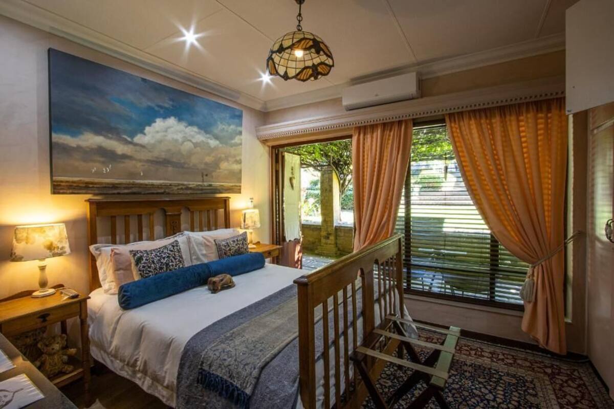 A bedroom with a bed, a nightstand, a lamp, a painting, a window with curtains, and a view of greenery outside.