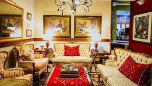 A traditional room with a sofa, armchairs, a red ottoman, a chandelier, framed pictures, and a view of the outside.