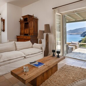 Una sala de estar con sofá, una mesa de centro de madera y vista al mar a través de un ventanal.