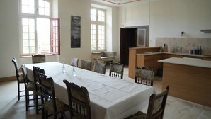 Uma área de refeições com mesa posta para seis pessoas, rodeada por cadeiras, uma área de cozinha com bancada e uma grande janela com vários painéis.