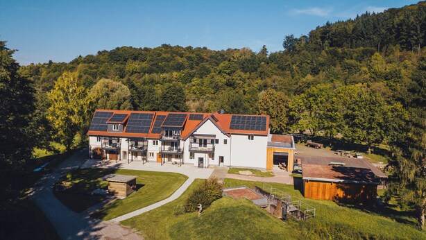 Ein mehrstöckiges Gebäude mit Solaranlage auf dem Dach, umgeben von üppiger Vegetation und einem bewaldeten Hügel.