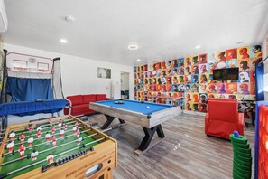 Una habitación con mesa de futbolín, mesa de billar, sofá rojo y una pared decorada con arte pop.