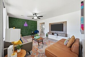 Una sala de estar moderna con una pared verde, un ventilador de techo, un televisor de pantalla plana, un sofá, una mesa de centro y una alfombra estampada.