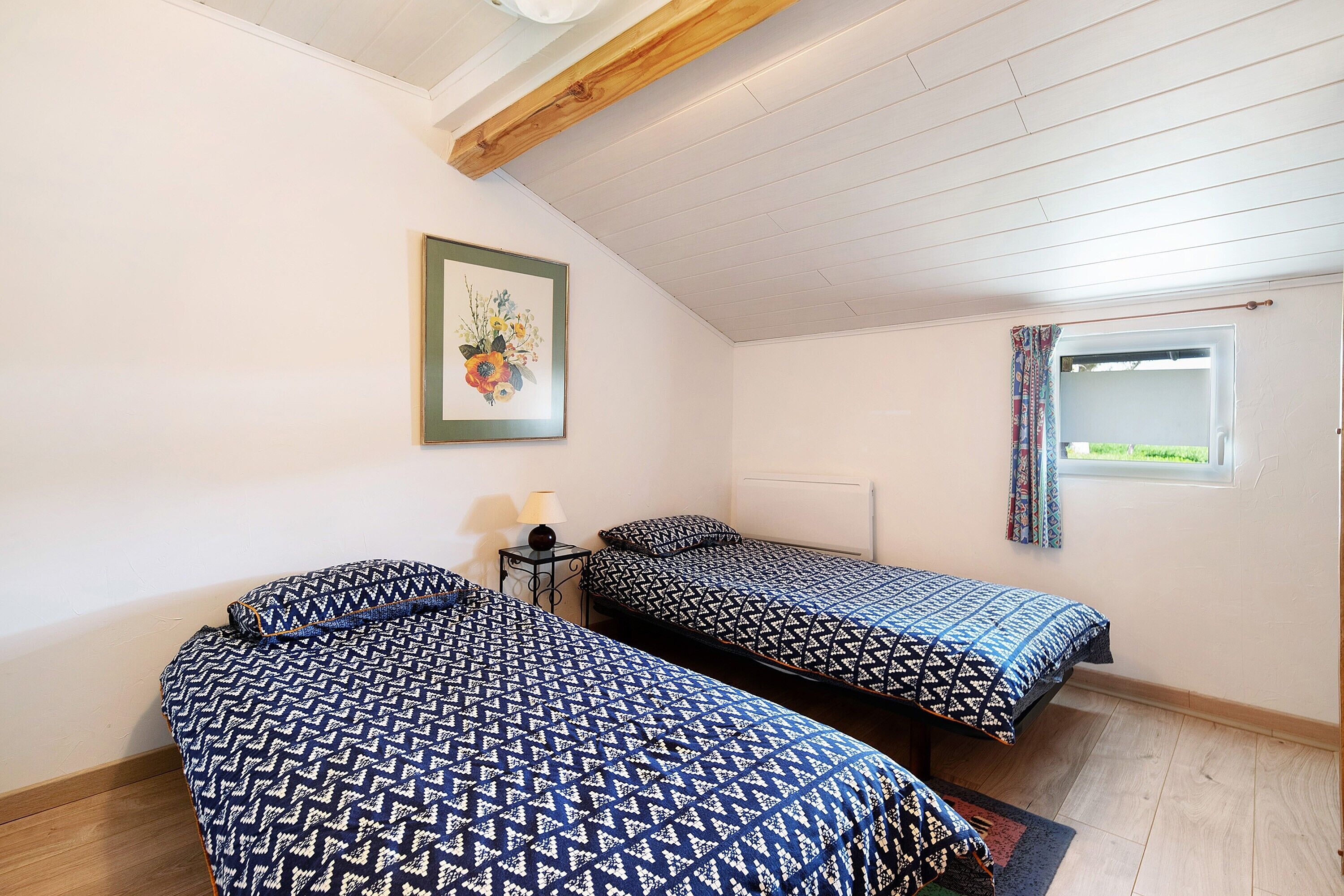 Een kamer met twee bedden, een ingelijst kunstwerk, een raam met gordijnen en een tafeltje met een lamp.