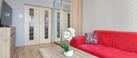 A modern living room with a red sofa, a wooden coffee table with a potted orchid, a TV on a wooden cabinet, and a mirrored wardrobe.