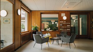 Aula, jossa on puinen vastaanottotiski, kaksi harmaata nojatuolia, pieni keltainen pöytä ja ruukussa oleva kasvi.