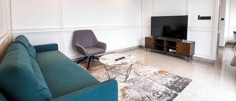 A modern living room with a teal sofa, a grey armchair, a coffee table, and a flat-screen TV on a wooden stand.