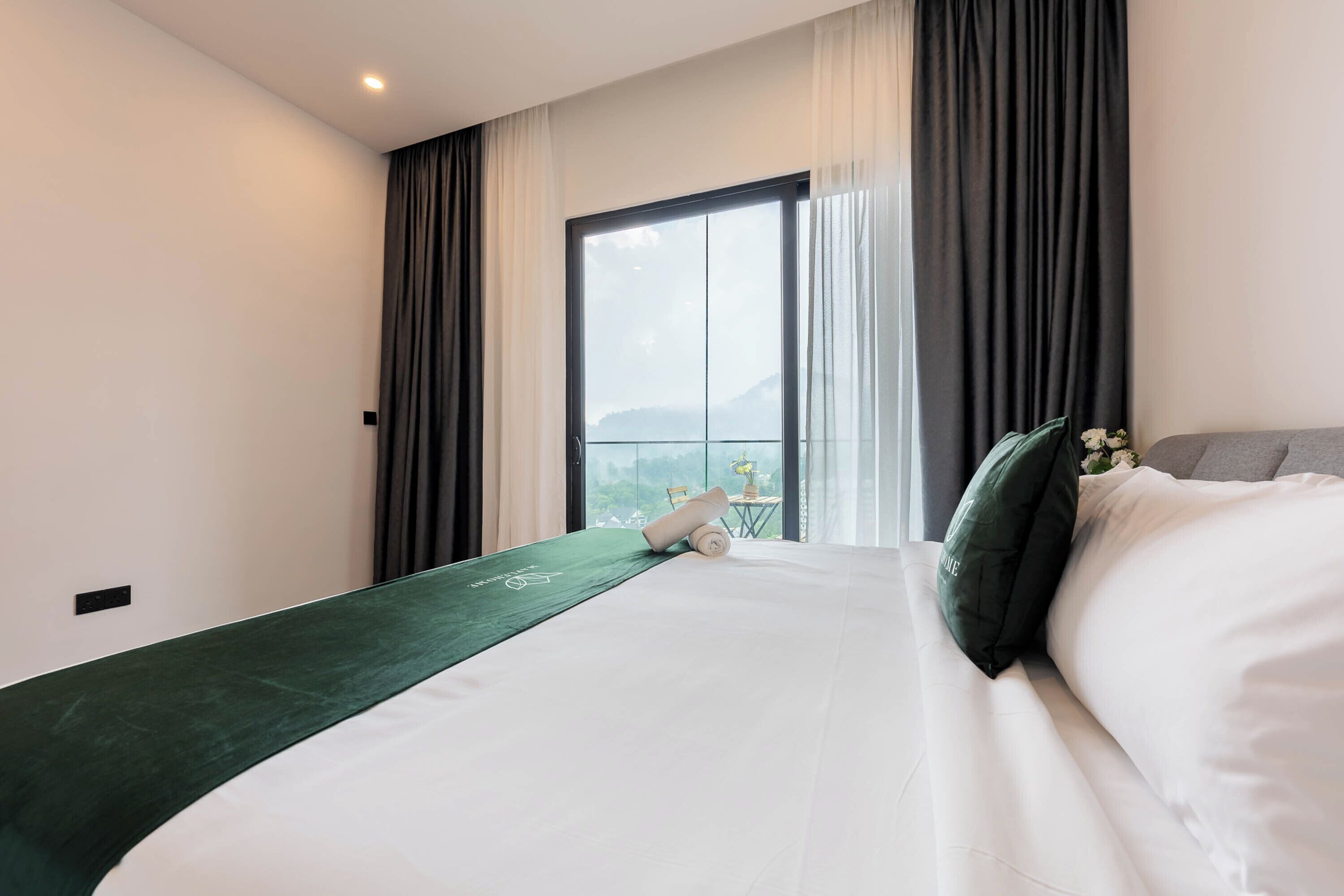 A hotel room with a large bed, a green runner, a view of a mountain through a window, and a small table with a plant.
