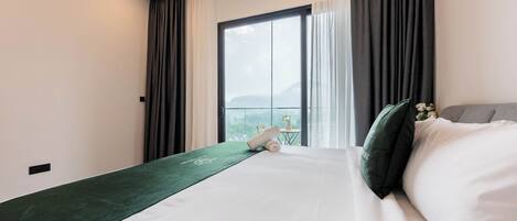 A hotel room with a large bed, a green runner, a view of a mountain through a window, and a small table with a plant.
