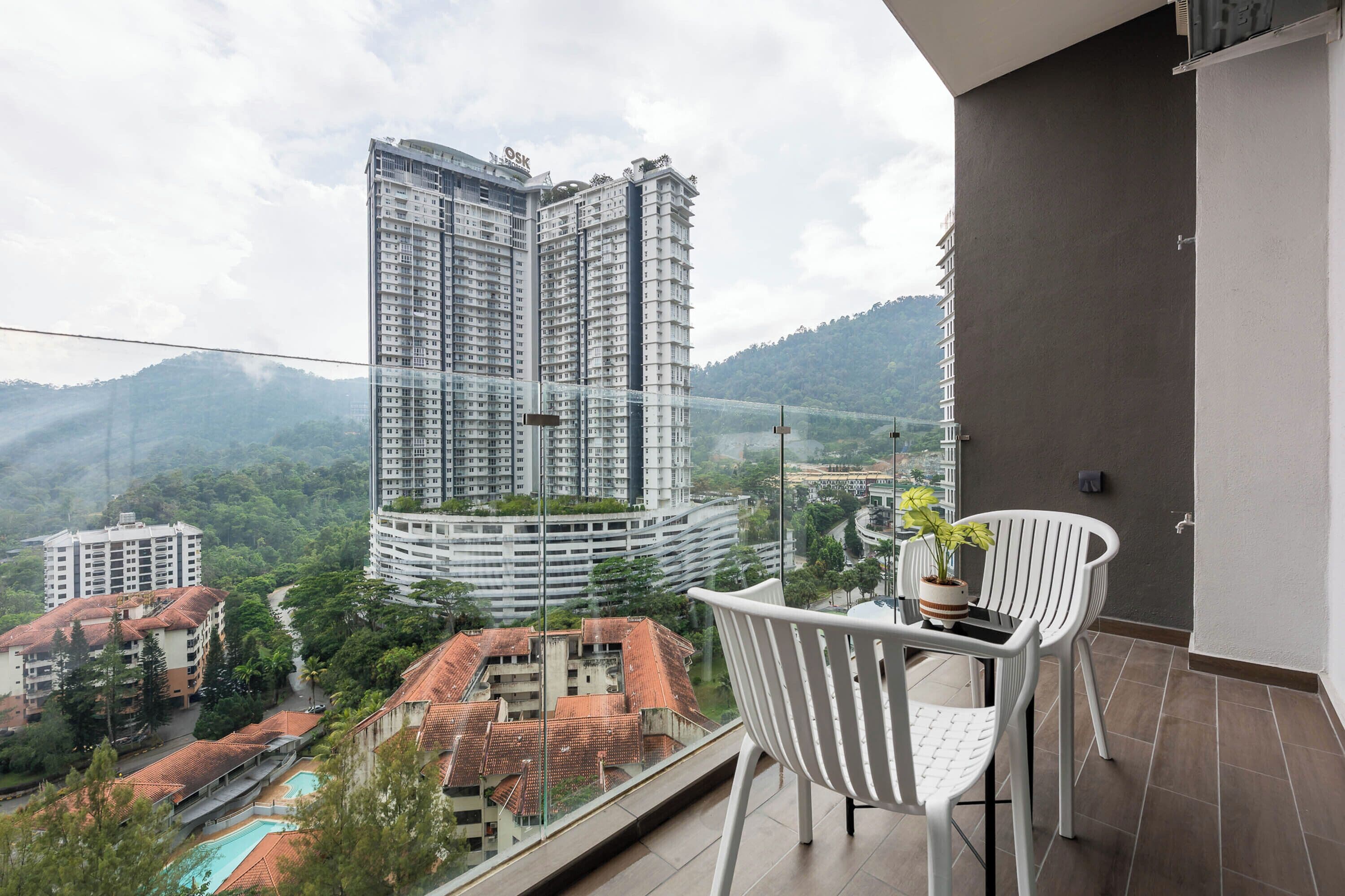 A balcony with a white chair and a table, overlooking a cityscape with high-rise buildings and a mountainous background.